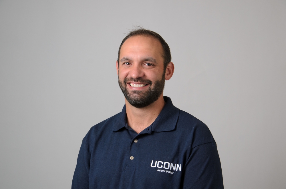 Portrait of Travis Beausoleil wearing a blue collared shirt with UConn Avery Point logo